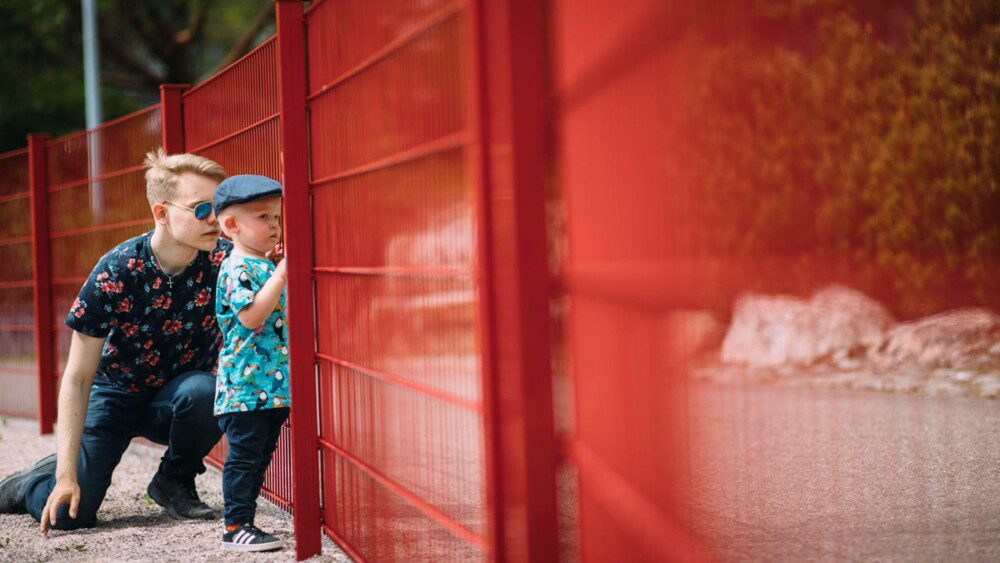Nuori ja lapsi katsovat punaisen aidan läpi leikkipuistoon toiselle puolelle.