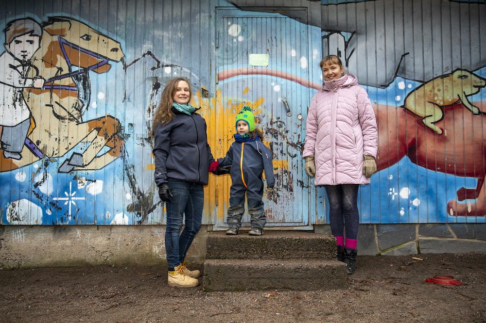 Kaksi naista seisovat pienen pojan kanssa ulkona graffiteilla maalatun seinän edustalla.